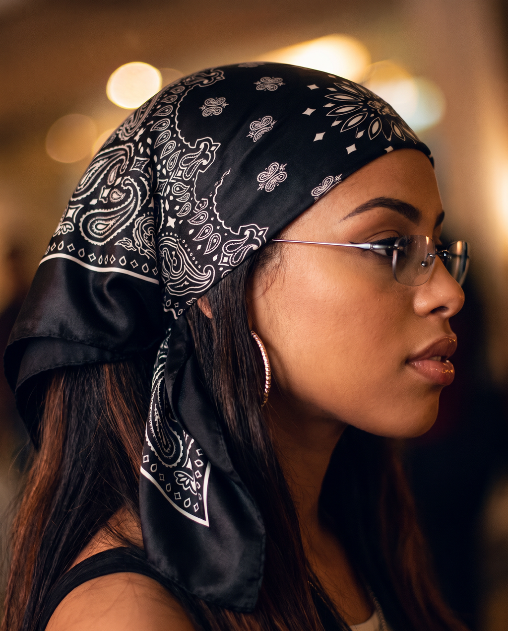 A woman in profile wearing a black silk bandana with a white paisley pattern, styled as a headscarf. She is wearing thin-framed glasses and hoop earrings against a warm, blurred background.